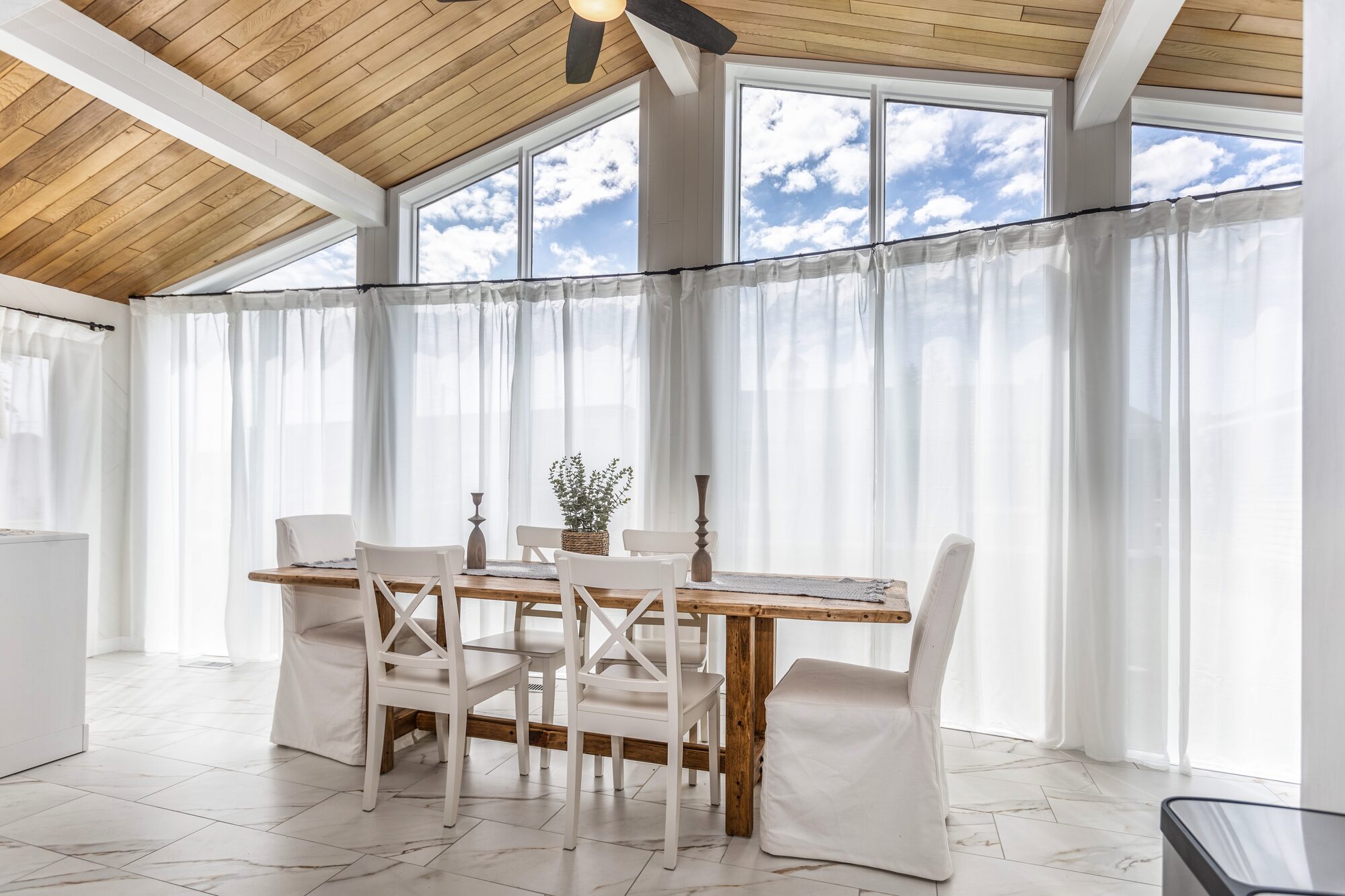 The sun-filled dining room with vaulted wood ceiling and tall windows draped in white sheers.