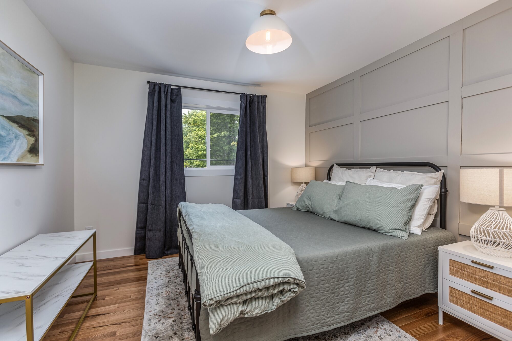 The downstairs queen bedroom with sage-green linen, a panelled accent wall, and a coastal painting.